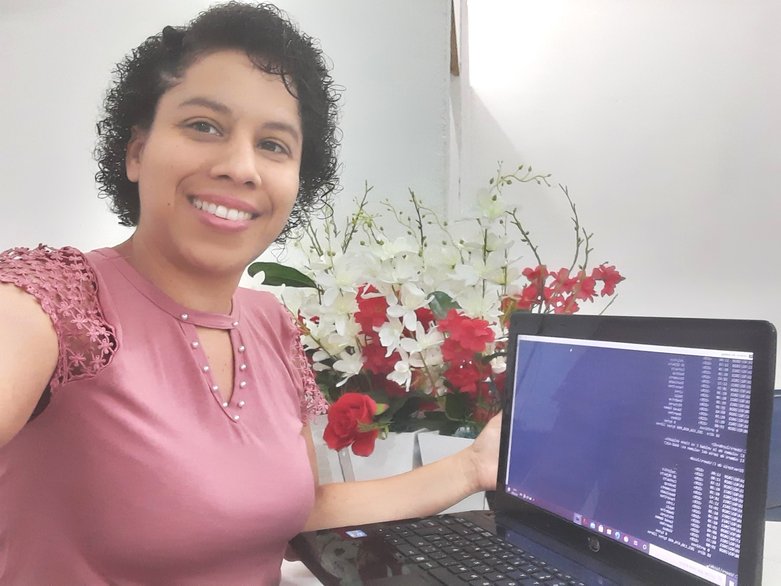 Luzdalis é uma pessoa do sexo feminino, parda e venezuelana. Na imagem, ela está sentada em frente a um notebook que exibe uma tela com códigos. Ela olha para a câmera e sorri.