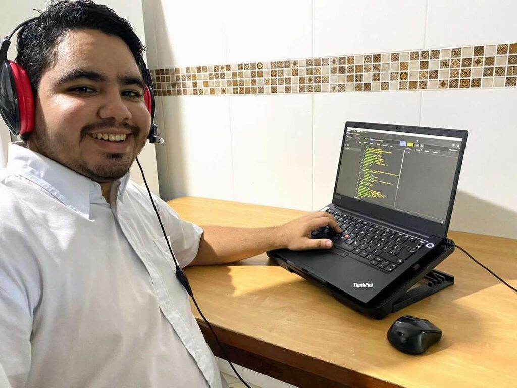 Wendell é uma pessoa do sexo masculino, branco e venezuelano. Na imagem, ele está sentado em frente a um notebook que exibe uma tela com códigos. Ele olha para a câmera e sorri. 
