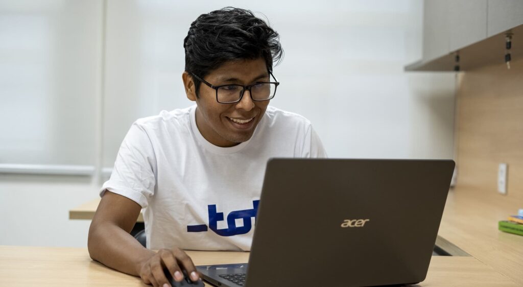 Profissional formado pela Toti como desenvolvedor full stack está sentado em frente a um notebook. Ele é boliviano, pardo e usa óculos de grau