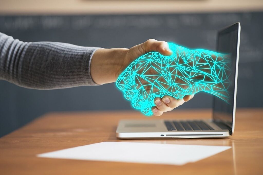 Imagem de um aperto de mão entre uma pessoa e uma espécie de robô, que está saindo de um notebook. A foto transmite uma ideia de interação entre seres humanos e inteligência artificial.