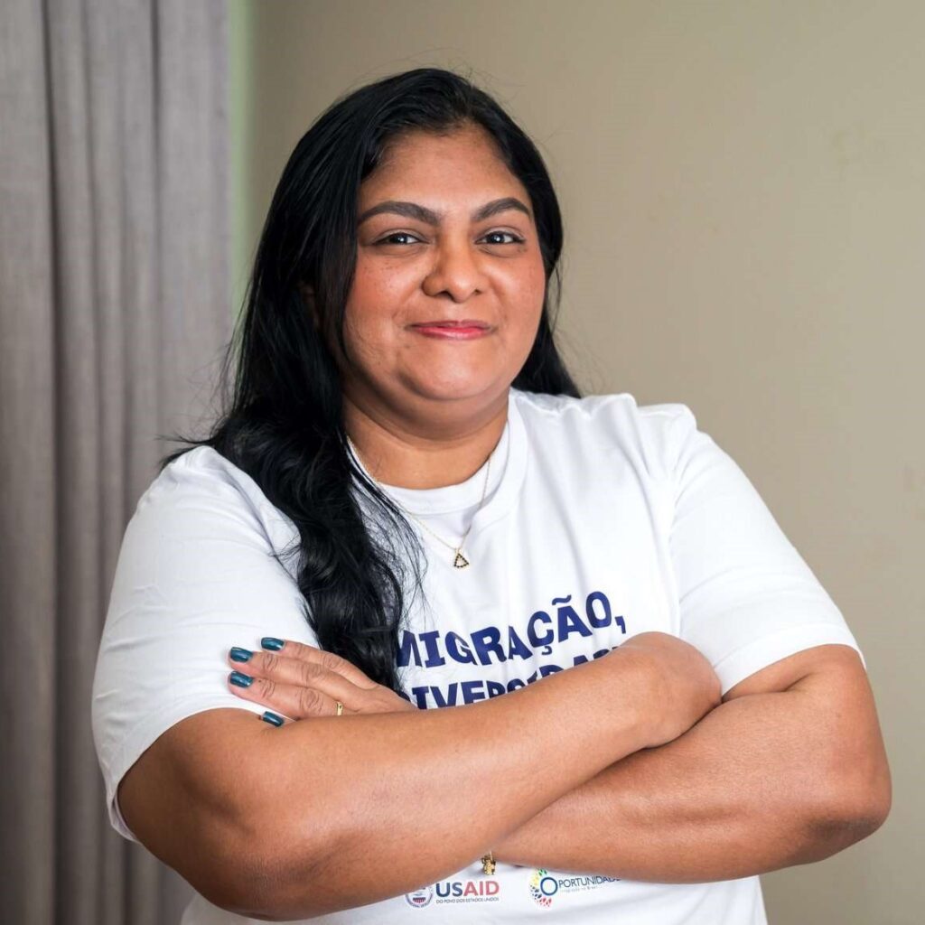 Marianela está em pé e veste uma camisa branca escrito "Migração, diversidade e tecnologia". Ela é venezuelana, possui cabelo preto e é parda.